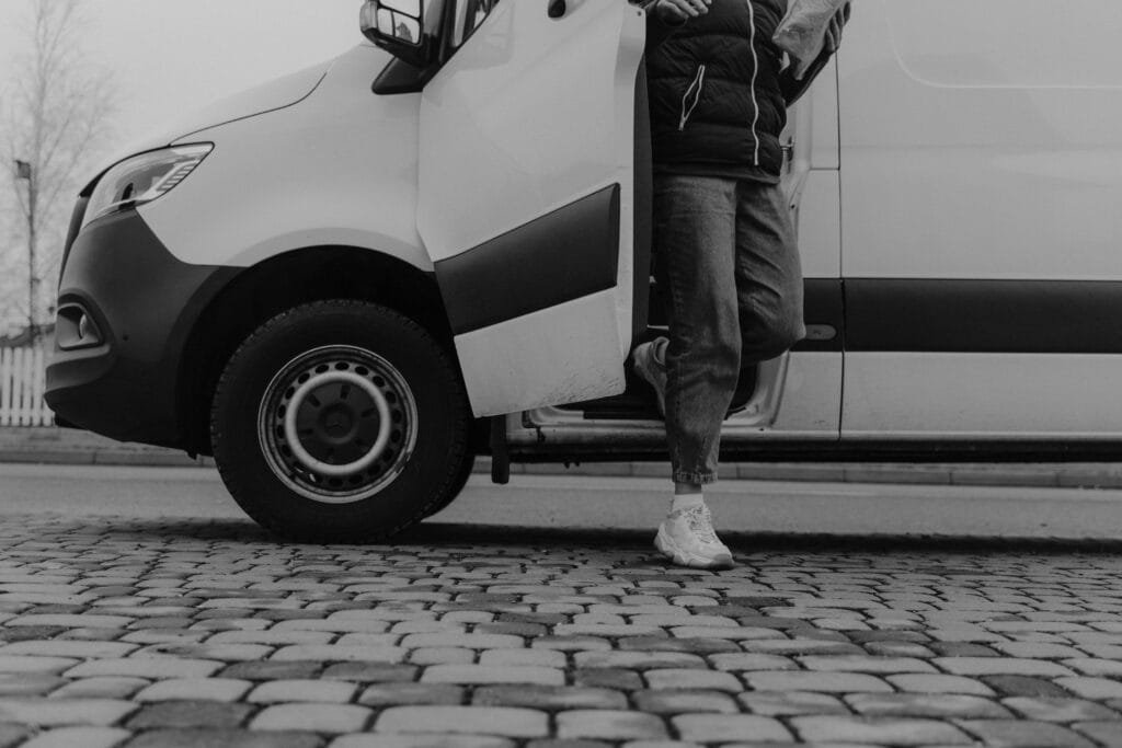 Monochrome image of a person stepping out of a van onto cobblestone pavement.